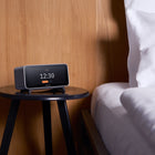 Digital clock on a wooden side table next to a bed with white bedding.