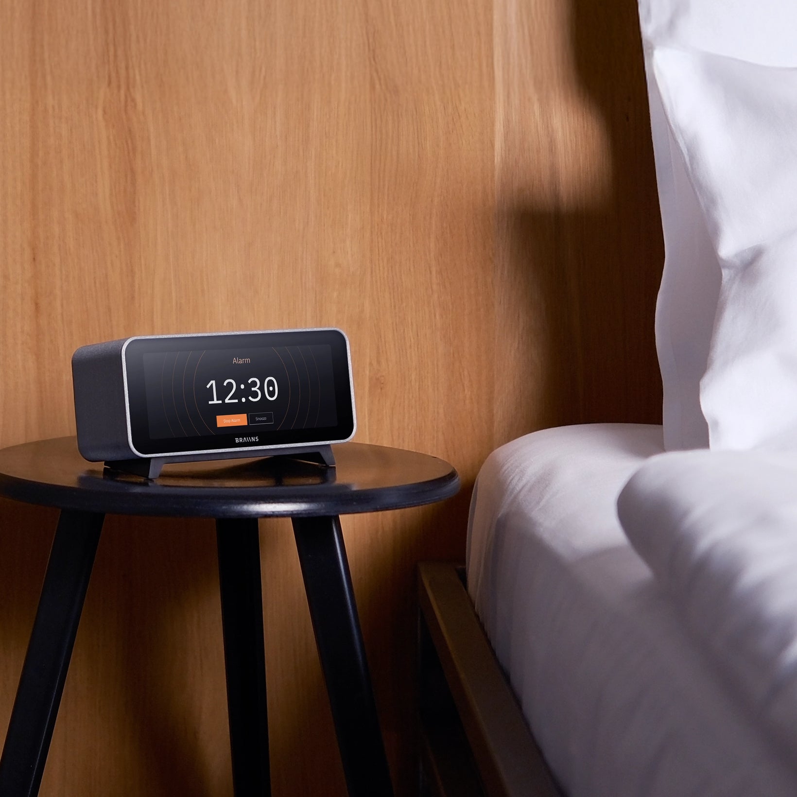 Digital clock on a wooden side table next to a bed with white bedding.